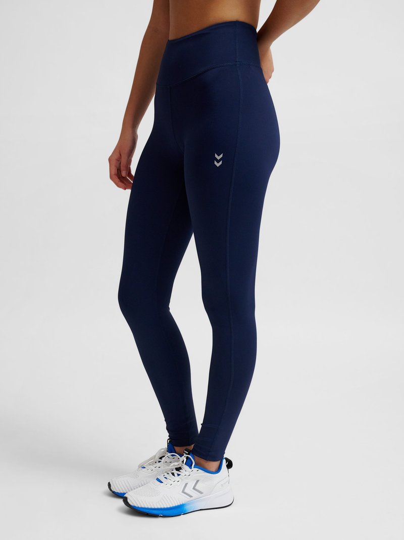 Person trägt marineblaue Leggings mit hoher Taille und weiße Sportschuhe mit blauen Akzenten, steht vor einem schlichten Hintergrund.