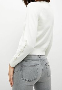 Biały sweter z dzianiny z długimi rękawami, ściągaczami na mankietach oraz haftem "Liu Jo" na rękawie. W połączeniu z dopasowanymi szarymi dżinsami.