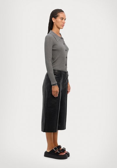 Frau mit geflochtenem Haar steht seitlich und trägt einen grauen Knopf-Pullover, schwarze strukturierte Culottes und schwarze Plateausandalen mit Schnallen.