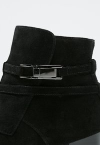 Bottine en suède noire avec une texture lisse, présentant un détail de boucle métallique et des accents de couture sur le côté.