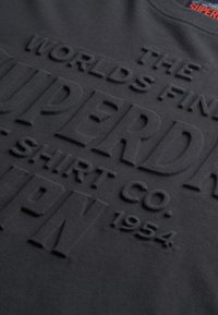 Sort bomulds t-shirt med hævet, tekstureret skrift, der har "SUPERDRY" og "SHIRT CO." i fremtrædende, fed skrift. Blød stofoverflade.