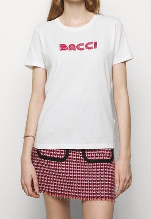 Woman wearing a white T-shirt with red "BACCI" text and a pink and black houndstooth mini skirt with front pockets.
