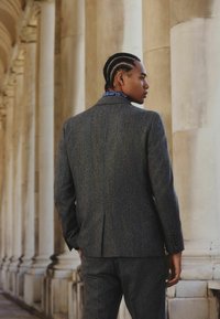 Gray tweed suit with a tailored fit, featuring a notched collar and buttoned cuffs. Blue patterned scarf visible at the neck.
