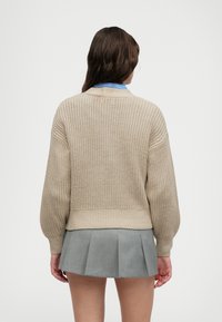 Lys beige strikket genser med løs passform og ribbet kant, kombinert med en grå plisserte skjørt. Bakfra vises det teksturerte strikkemønsteret.