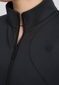 Giacca atletica nera con colletto alto, chiusura zip frontale, tessuto texturizzato e discreta dettagli del logo. Presenta cuciture tono su tono e un design elegante.