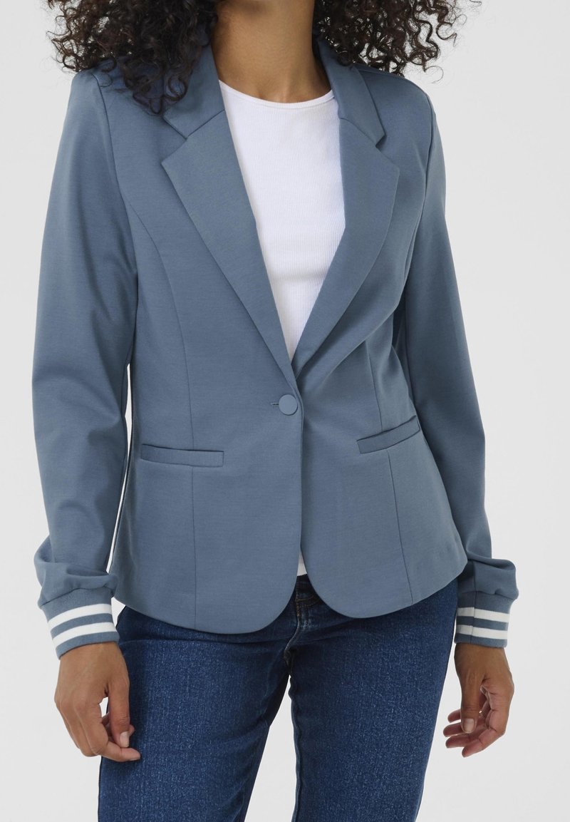Femme portant un blazer bleu à un bouton avec des poignets rayés sur une chemise blanche et un jean bleu, debout devant un fond uni.