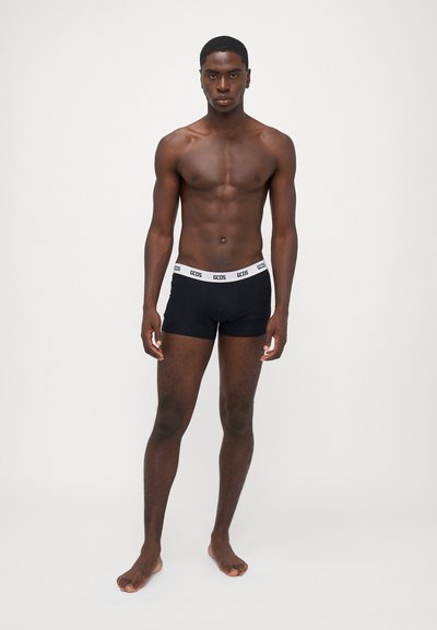 Schwarze Boxershorts mit einem weißen elastischen Bund, versehen mit dem Logo "GCOS." Das Modell steht barfuß mit einem neutralen Ausdruck vor einem weißen Hintergrund.