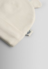 Krémovo sfarbená rebrovaná látka s okrúhlym tvarom a malými uškami. Obsahuje našitú štítkovú značku s číslom „90“ vo tmavomodrej farbe.