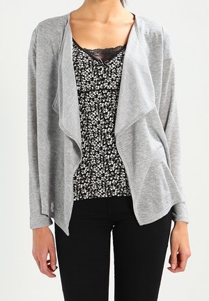 Vrouw draagt een lichtgrijs openvallend vest over een zwarte bloemenblouse met kanten rand, gecombineerd met een zwarte broek tegen een effen achtergrond.