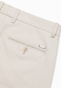 Tasca posteriore di pantaloni beige con chiusura a bottone, passanti per cintura e piccola targhetta rettangolare in metallo del marchio su tessuto strutturato.