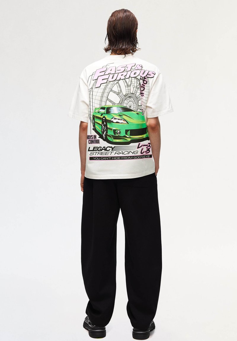 Person with shoulder-length hair wearing white Fast & Furious T-shirt with green sports car and tire graphic, paired with black wide pants and shoes.