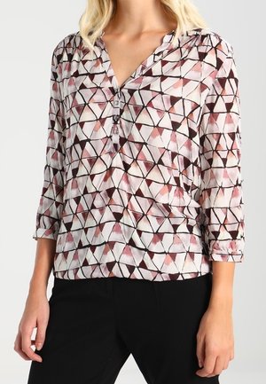 Woman wearing a long-sleeve blouse with abstract pink, purple, and black triangular pattern and three-button V-neck paired with black pants.
