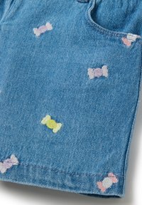 Pantalones cortos de mezclilla con formas de caramelos bordadas multicolores en púrpura, rosa y verde sobre un fondo azul claro. Diseño clásico de cinco bolsillos.