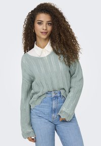 Maglione lavorato a maglia verde chiaro con una vestibilità ampia e un orlo increspato, indossato sopra una camicia bianca con colletto. Presenta maniche lunghe e larghe.