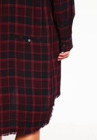 Robe chemise à manches longues à carreaux rouge et noir, avec poignets à boutons, poche arrière avec fermeture à bouton, et ourlet à franges.