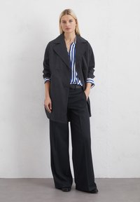 Palton negru cu două rânduri de nasturi și guler lat, purtat cu o cămașă cu dungi albastre și albe și pantaloni negri lejeri. Textura fină a materialului.