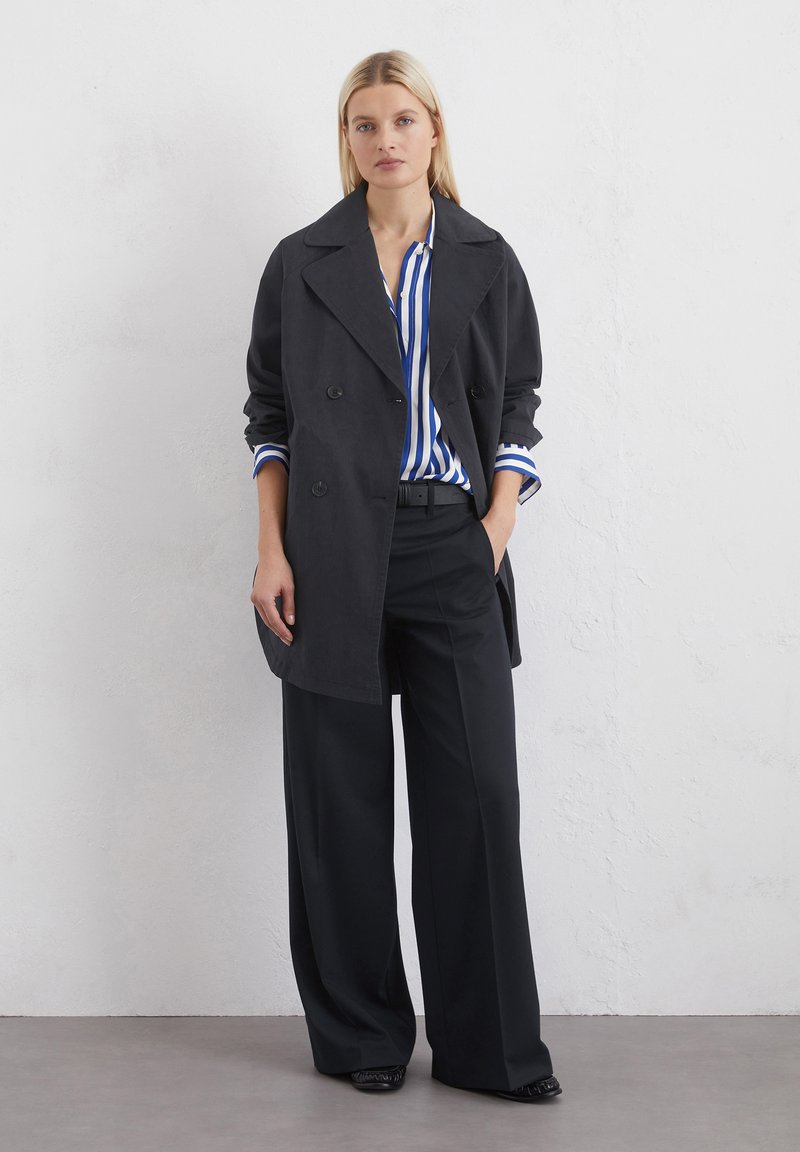 Czarny dwurzędowy płaszcz trench z szerokim kołnierzem, zestawiony z niebiesko-białą w paski koszulą i luźnymi czarnymi spodniami. Gładka tekstura tkaniny.