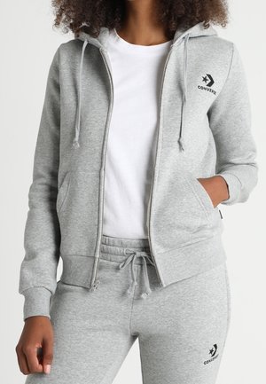 Femme portant un sweat à capuche gris clair zippé et un pantalon de survêtement assorti avec des logos Converse noirs, un T-shirt blanc en dessous, main dans la poche.