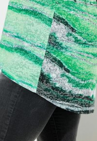 Zielono-szara bluzka z falistym wzorem, wykonana z miękkiego materiału. Charakteryzuje się zaokrąglonym dołem i luźnym krojem, zestawiona z ciemnymi dżinsami.