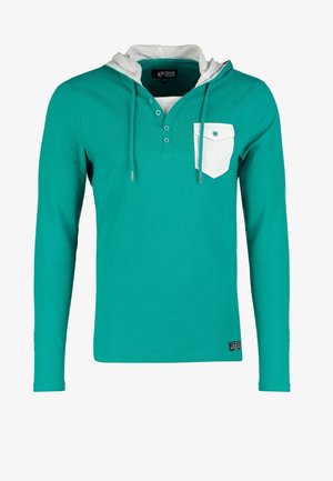 Mężczyzna idzie w tealowej bluzie z kapturem z białą kieszenią, ciemnych dżinsach, białych sneakersach i nosi czarną torbę na ramię.