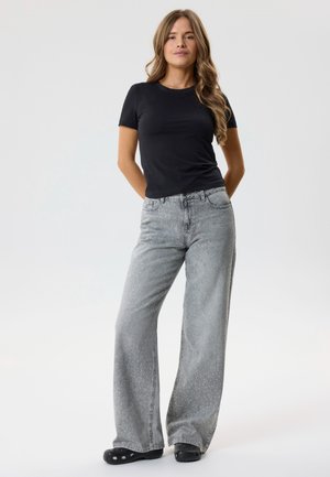 Kobieta z długimi, falującymi włosami stoi, ubrana w czarną koszulkę z krótkim rękawem, jasnoszare, szerokie spodnie z małymi kropkami oraz czarne buty wsuwane.