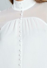 Blusa blanca con un cuello alto, que presenta una tapeta frontal con seis botones redondos y detalles plisados, hecha de una tela sheer y texturizada.