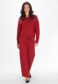 Burgundské pletené svetr s V-výstřihem a flitrovým designem na ramenou, kombinovaný s volnými kalhotami v odpovídající barvě, vytvářející soudržný outfit.