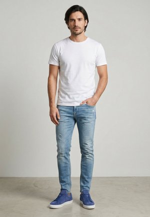 Man staat met één hand in zijn zak, draagt een wit T-shirt, lichtblauwe spijkerbroek en blauwe sneakers tegen een effen achtergrond.