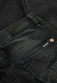 Jeans de mezclilla negra con una etiqueta plana en la cinturilla, bolsillos delanteros y traseros, y costuras en contraste. Brillo sutil en la tela.