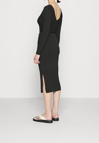 Robe noire côtelée avec des manches longues et un dos en V, présentant une silhouette ajustée et une fente sur le côté. Portée avec des sandales blanches à glisser.