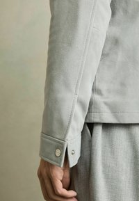 Mâneca unei jachete din suede gri, cu un manșon cu nasture prin presare, textură netedă și croială ajustată, combinată cu un articol vestimentar din textil de culoare gri deschis dedesubt.