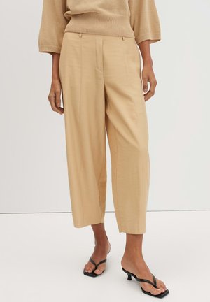 Model trägt beige weite, knöchellange Hose, beigen Pullover und schwarze Sandalen mit dünnen Riemchen und Absatz vor einem einfarbigen hellen Hintergrund.
