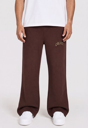 Mand iført brune bukser med vide ben med teksten "COLUCCI" på venstre lår og hvide sneakers mod en ensfarvet baggrund.