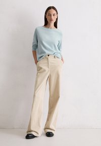 Helles blaues Langarmoberteil mit rundem Ausschnitt. Beige Corduroy-Hose mit weitem Bein, vertikalen Streifen und Fronttaschen. Schwarze flache Schuhe.