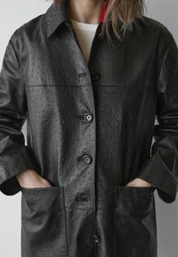 Manteau en cuir noir avec un motif texturé, équipé d'une fermeture à boutons, de deux poches avant et d'un col. L'intérieur présente une touche rouge.