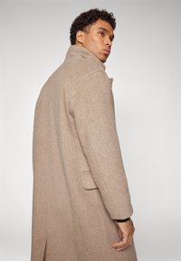 Beige Wollmantel mit hohem Kragen, tailliertem Schnitt und seitlichen Taschen, ausgestattet mit einer dezenten Textur und einem Schlitz hinten.