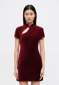Burgundowa welurowa sukienka z krótkimi rękawami, z wysokim kołnierzem, wycięciem w kształcie klucza oraz dekoracyjnym węzłem przy dekolcie.