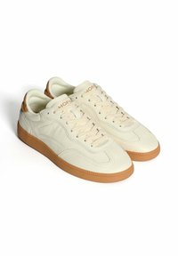 Zapatillas de cuero crema con suelas de goma, cierre de cordones y cuellos acolchados, posicionadas una al lado de la otra sobre un fondo blanco.