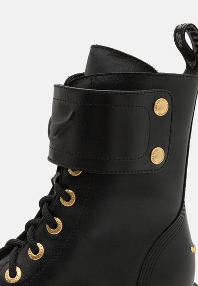 Svart läderankelboots med detaljer i guld, inklusive runda öglor och ett metallknäppt lås mitt på skon, strukturerad yta.