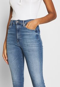 Jeans de denim azul de cintura alta con un ligero desvanecimiento, que cuentan con bolsillos frontales, cierre de botón y un diseño ajustado.