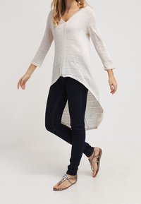 Lehký krémově pruhovaný top s výstřihem do V, tříčtvrtečními rukávy a asymetrickým lemem, spojený s tmavými úzkými džínami a hnědými sandály.