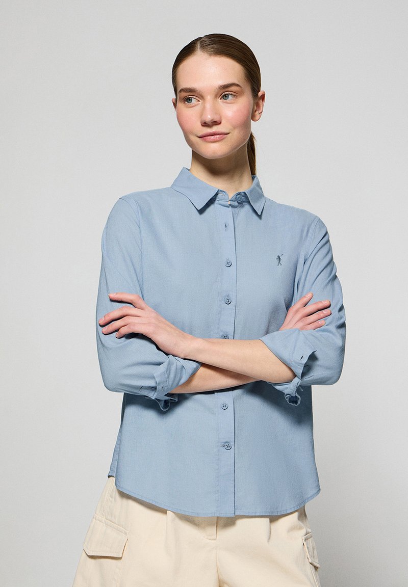 Camisa azul clara de botones con una textura suave, que presenta un cuello clásico, mangas largas remangadas y un pequeño logotipo en el pecho.