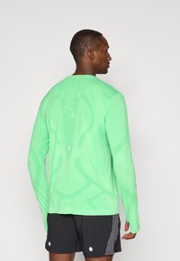 Langermet, lys grønn treningsskjorte med teksturert stoff og subtile designdetaljer bak, kombinert med svarte shorts.