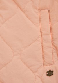 Prešito blago v mehki breskovi barvi z valovitim vzorcem šivov. Vsebuje majhen rjav gumb v obliki cvetlice in detajl stranskega žepa.