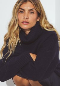 Tamsiai mėlynas turtlenecko megztinis, pagamintas iš minkštos, tekstūruotos medžiagos, su banguotais rankogaliais ir laisvu siluetu, turintis apykaklę, kuri sulankstyta.