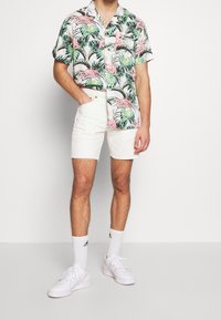 Camicetta a maniche corte con stampa tropicale di fenicotteri rosa, abbinata a pantaloni corti di jeans bianchi. Indossata con sneakers bianche e calzini alla caviglia.