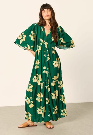 Kvinde med langt mørkt hår iført en grøn maxikjole med gult blomstermønster og vide ærmer, stående med hænderne på hofterne, brune sandaler.