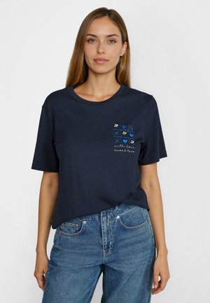 Frau trägt ein marineblaues T-Shirt mit einem kleinen, auf der linken Brustseite eingestickten Tic-Tac-Toe-Spiel und Text, kombiniert mit blauen Jeans.
