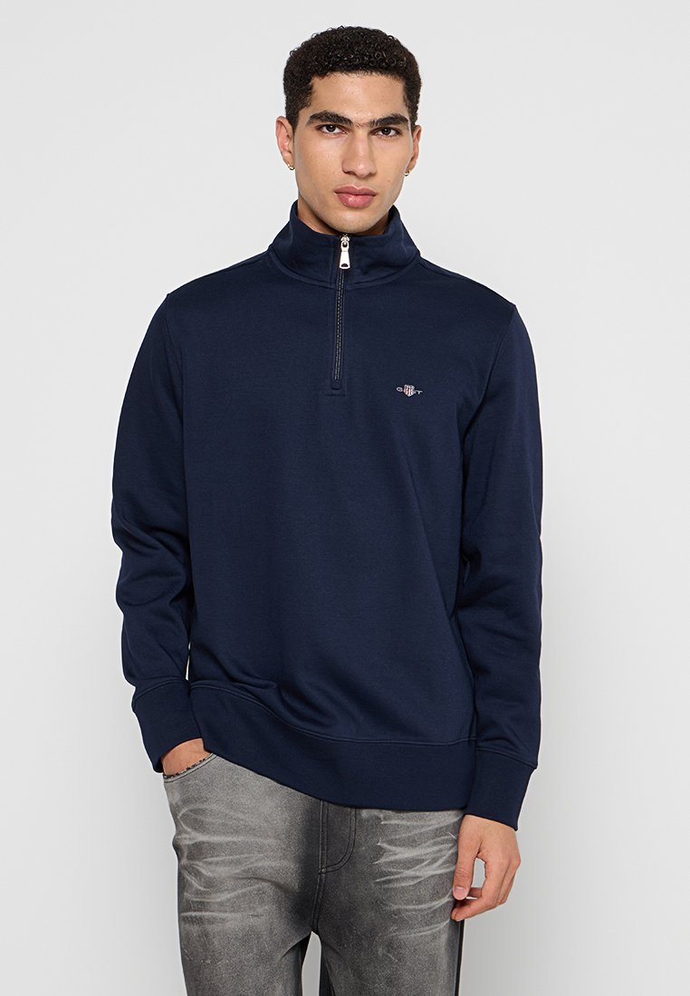Junger Mann in einem navyblauen Quarter-Zip-Pullover und grauen, ausgewaschenen Jeans, der mit einer Hand in der Tasche vor einem schlichten Hintergrund steht.