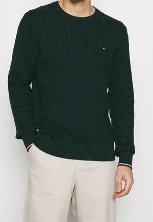 Mężczyzna w ciemnozielonym swetrze w grubym warkoczu oraz beżowych spodniach, z rękami częściowo w kieszeniach, stojący na jasnym, jednolitym tle.